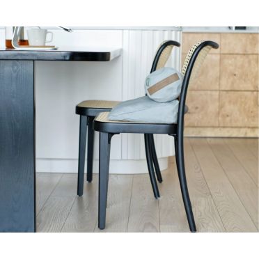 Mały wałek gryczany w kolorze jasnoniebieskim oraz dopasowana poduszka klinowa, ułożone na krześle z czarną ramą i plecionym oparciem, przy stole w domowej kuchni.